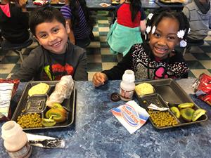 Two smiling elementary students sitting at a cafeteria table with their lunches set out before them. 